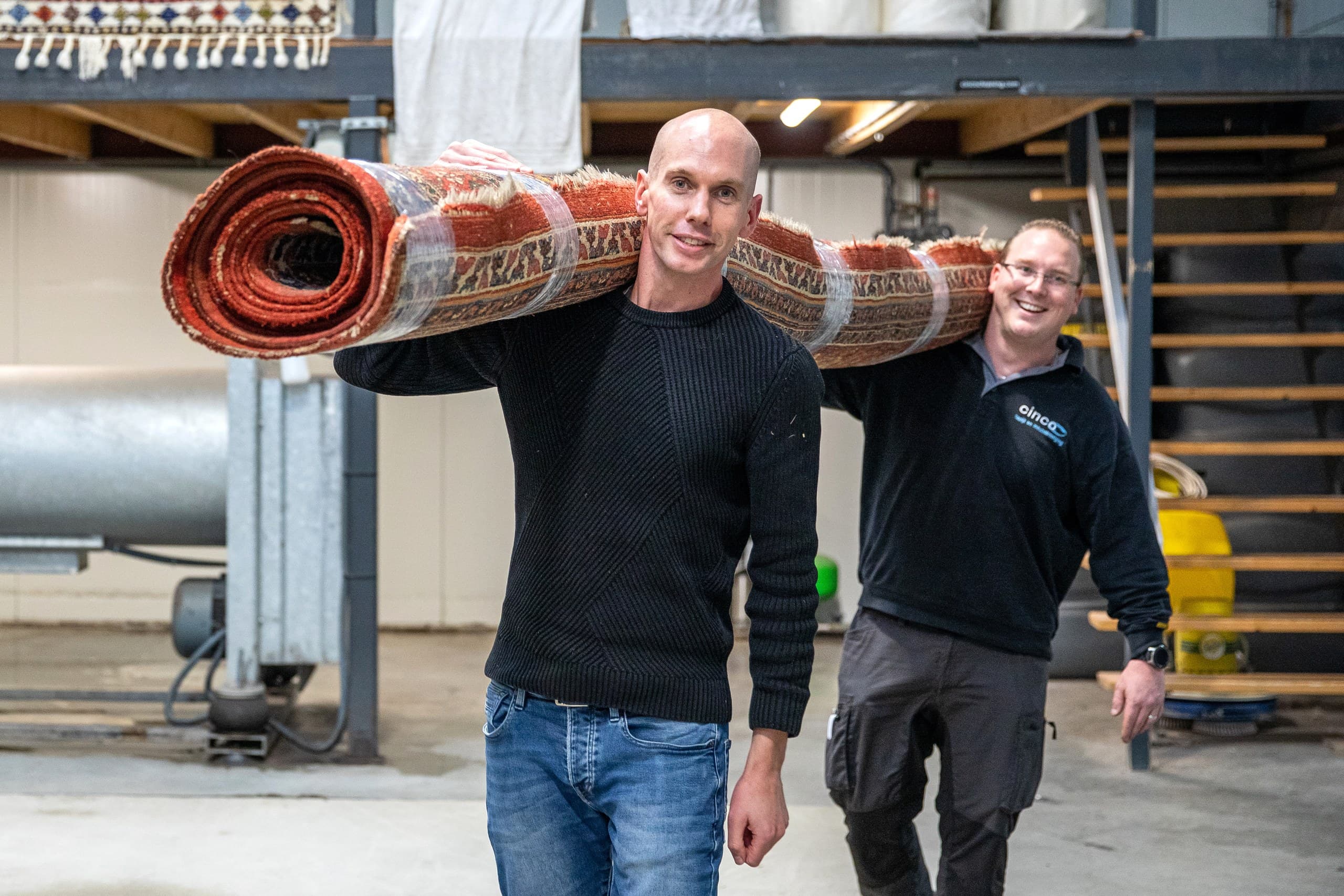 Twee medewerkers dragen een opgerold vloerkleed in de wasserij van Kleda